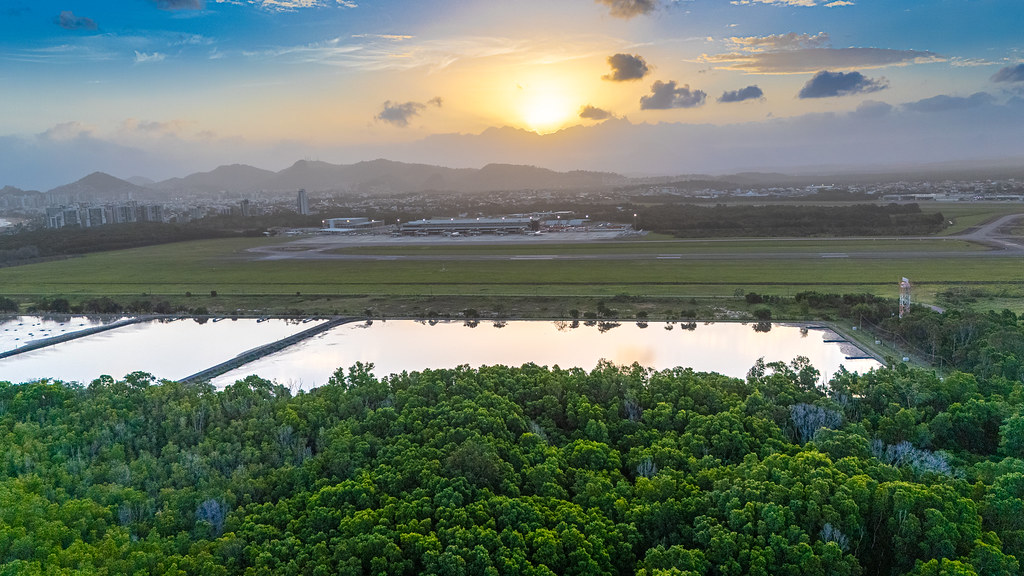 Foto aérea ao pôr do sol em Jardim Camburi, com vegetação, água, área urbana e montanhas no horizonte.